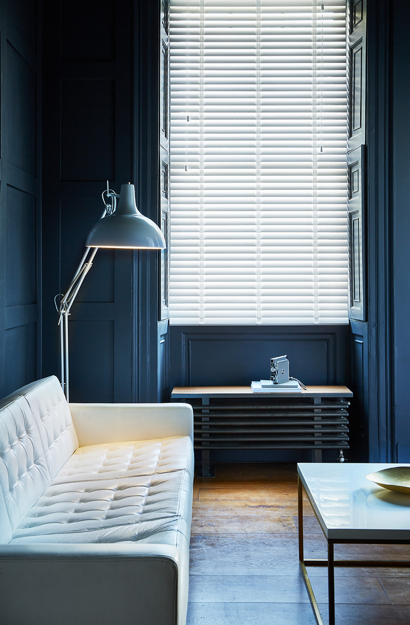 photo of dark blue themed room with white leather sofa next to window with white wooden blinds 