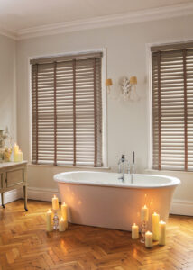 image of white stand alone bath with candles next to it in front of windows with brown faux wooden blinds