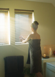 photo of woman standing in bathroom next to window with faux wooden blinds with sunlight shining through