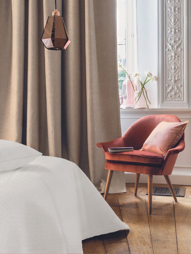 An orange chair by a window in a bedroom with a cream curtain